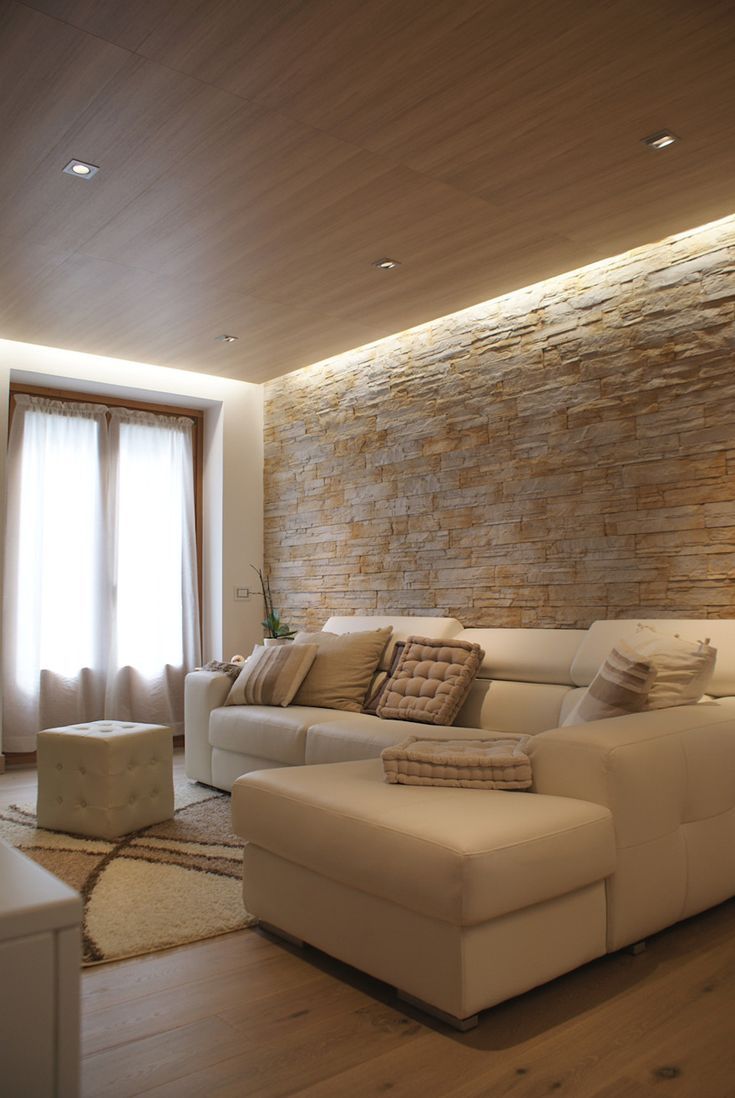 Accogliente soggiorno con divano componibile color crema, parete in pietra e tende trasparenti. Pavimento e soffitto in legno con illuminazione a incasso.
