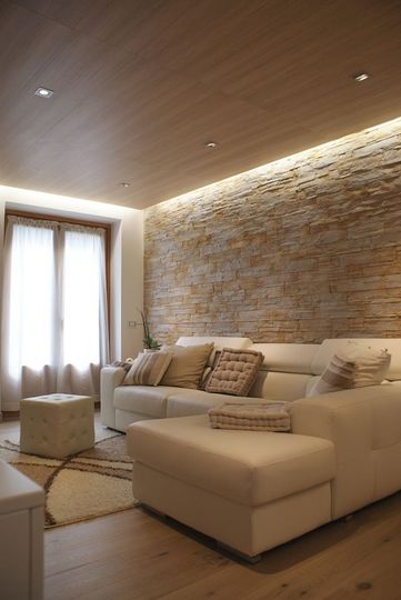 Accogliente soggiorno con divano componibile color crema, parete in pietra e tende trasparenti. Pavimento e soffitto in legno con illuminazione a incasso.