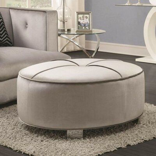 Pouf ovale grigio chiaro con finiture scure e piccole gambe argentate in un ambiente soggiorno. Sullo sfondo, un divano e un tavolino.