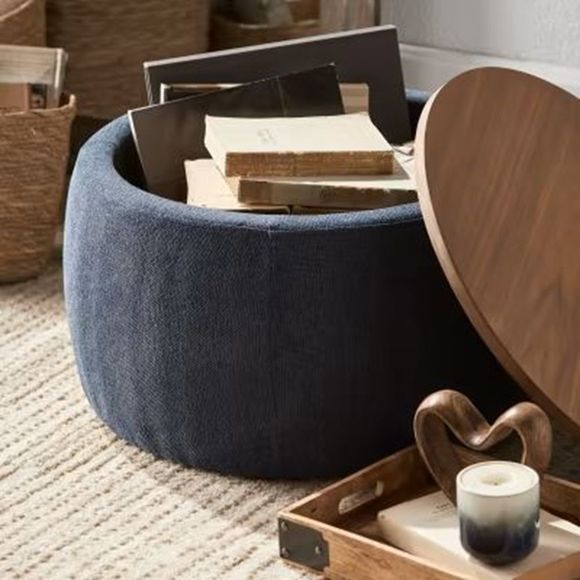 Un pouf contenitore rotondo blu navy con coperchio in legno, pieno di libri. È appoggiato su un tappeto color crema, con un vassoio e una scultura a forma di cuore nelle vicinanze.