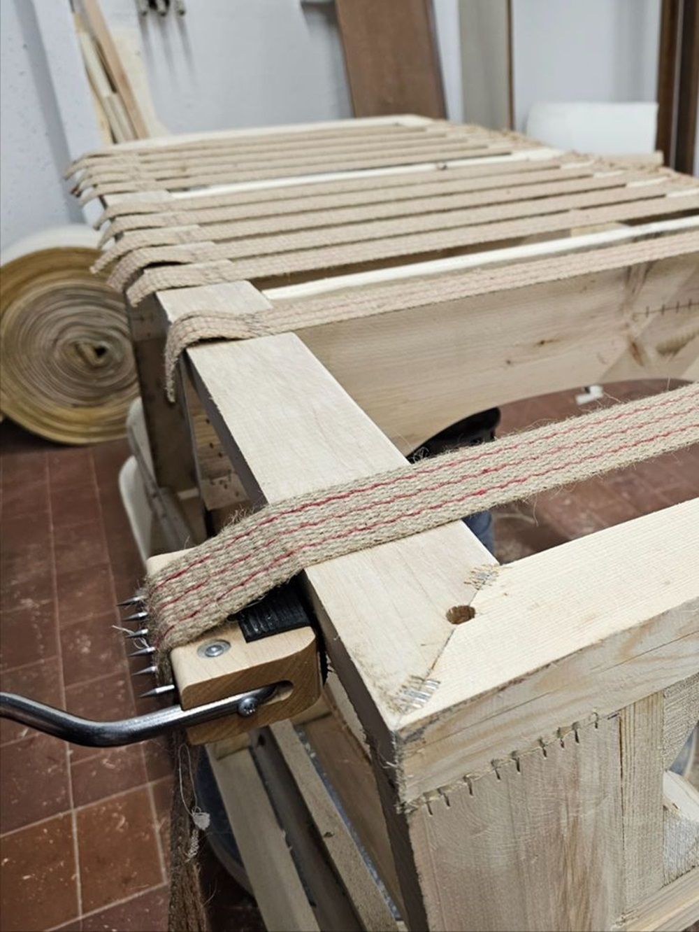 Un telaio di mobili in legno con cinghie tese tramite un tenditore. Sullo sfondo, un rotolo di tessuto.