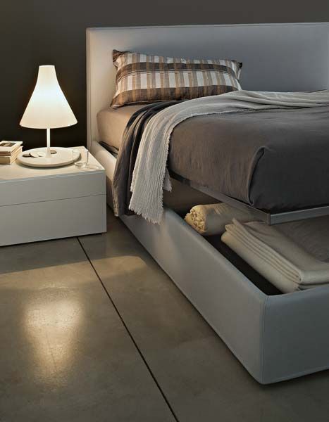 Camera da letto moderna con letto bianco, copriletto grigio e comodino con lampada. Un cassettone sotto il letto rivela la biancheria.
