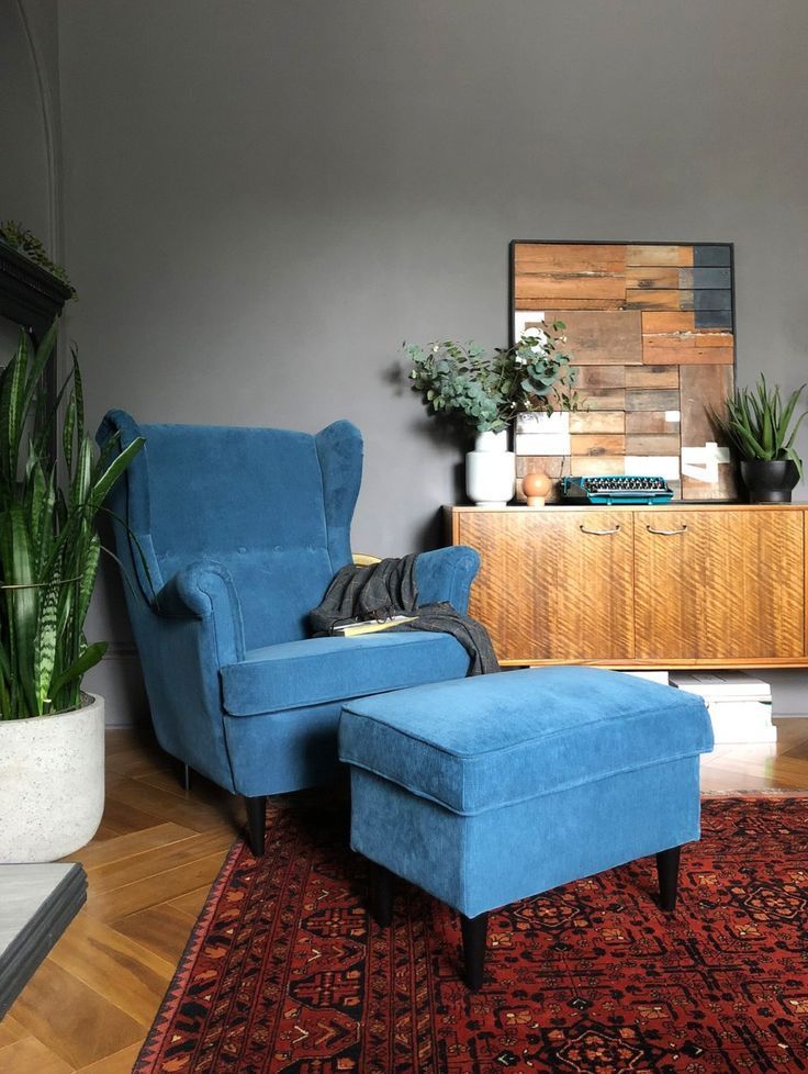 Una poltrona e un pouf blu sono appoggiati su un tappeto rosso a fantasia, accanto a un mobiletto in legno con opere d'arte e piante. Pareti grigie.