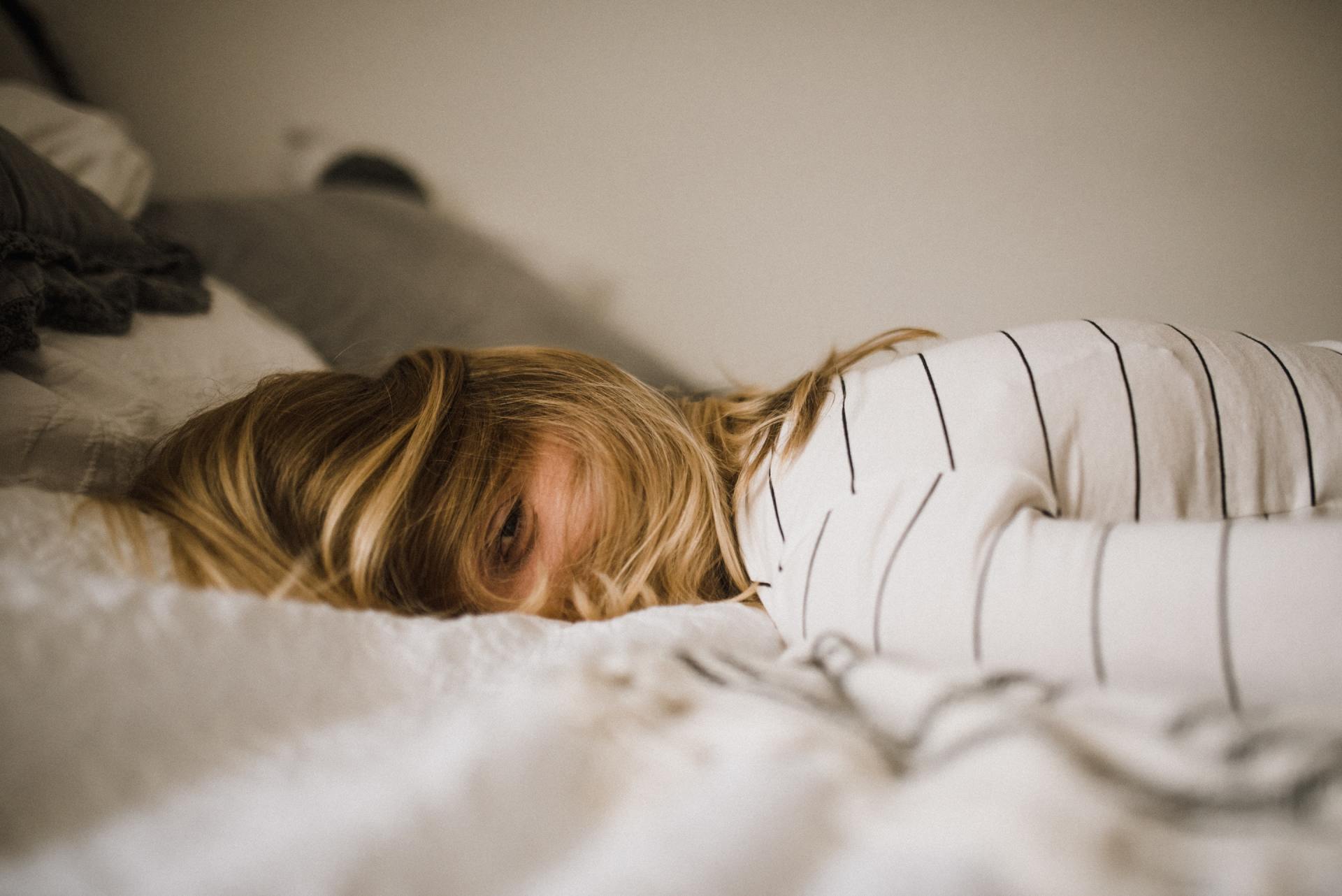 Person with blond hair lying face-down on a white bed, wearing a striped shirt, eyes open.