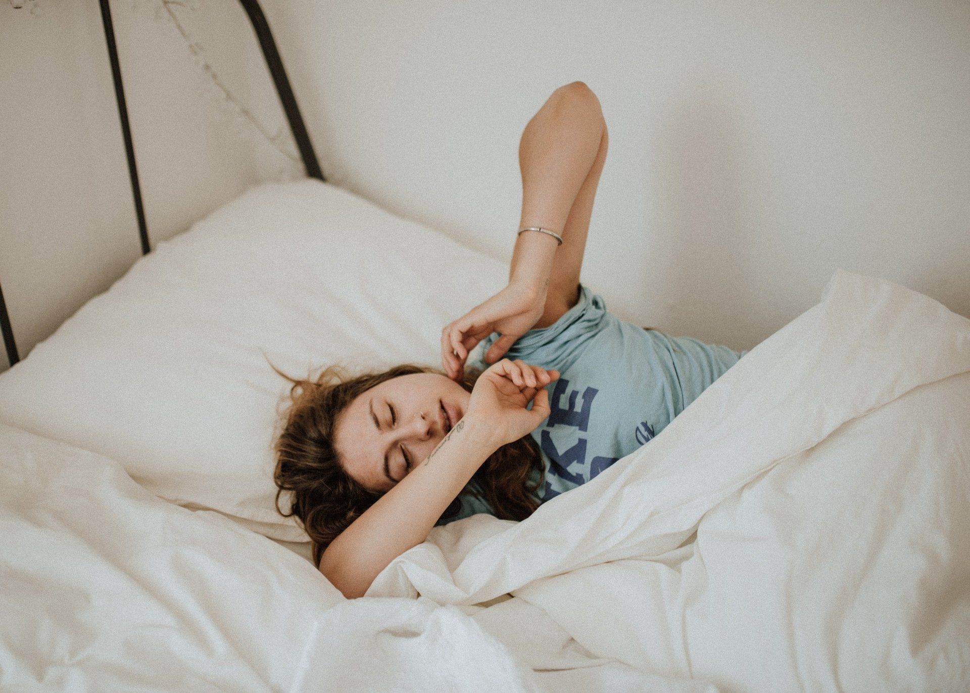 Woman waking up in bed, stretching arms, wearing a blue t-shirt, white bedding.