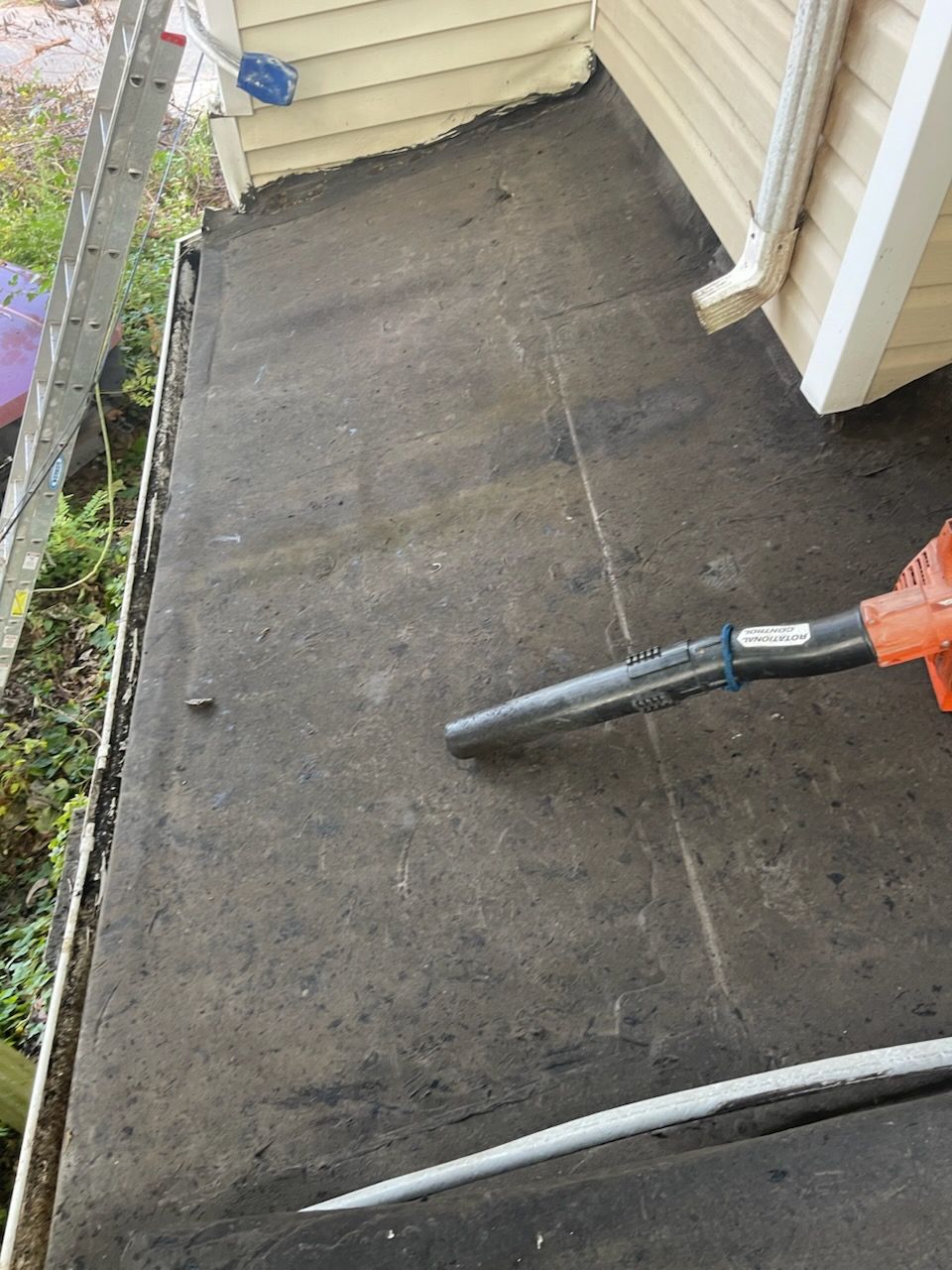 A person using a leaf blower on a flat, black roof near a light-colored building.