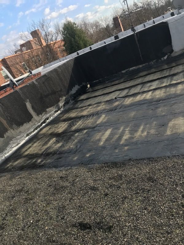 Flat, dark gravel roof; dark edging and water stain; cloudy sky and brick buildings in background.