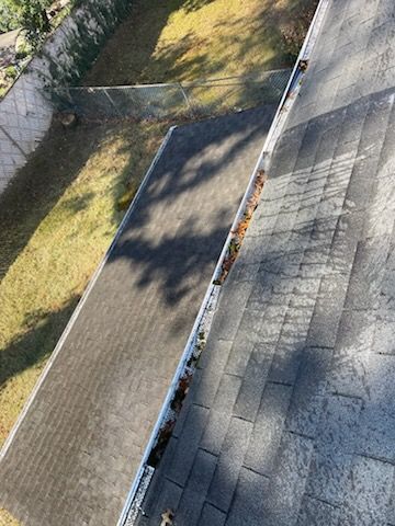 A rooftop with a gutter partially filled with leaves, overlooking a backyard with grass and trees.