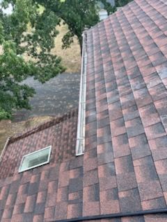 Angled view of a roof with brown and reddish shingles, a skylight, and a gutter, with trees in the background.