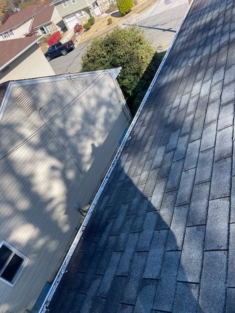 View from a roof of asphalt shingles. Next to it is a house with beige siding, a window, and a tree.