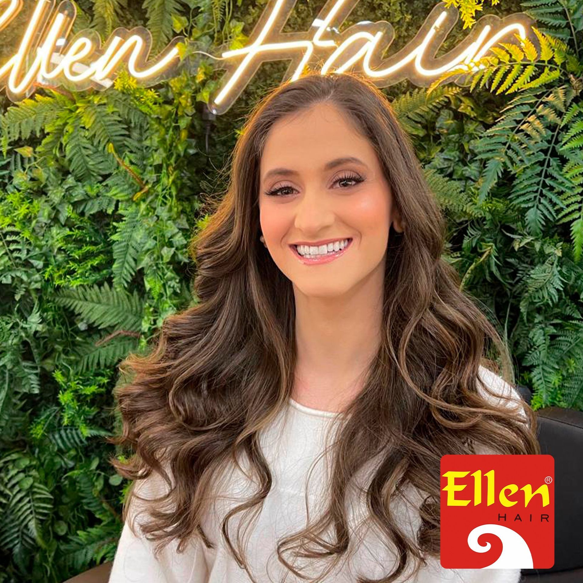 Mulher com longos cabelos castanhos ondulados sorrindo em frente a uma parede de plantas verdes e uma placa de neon.