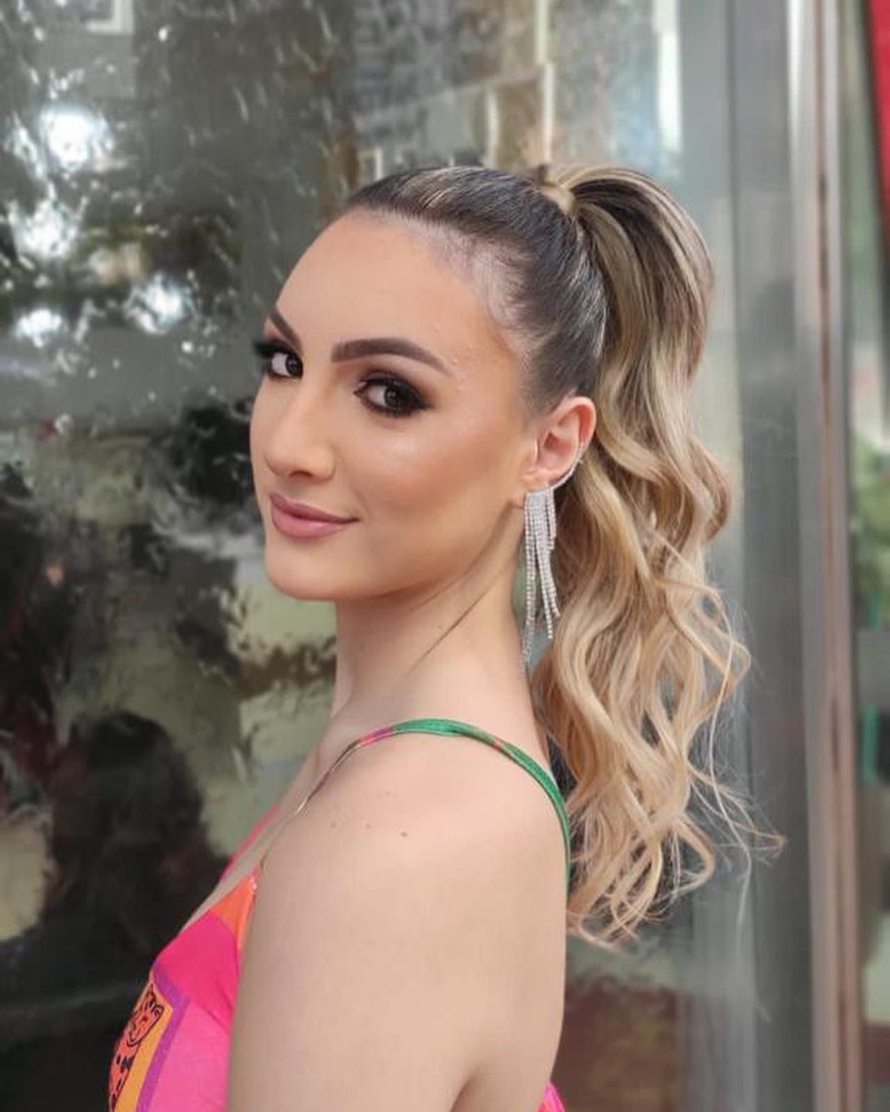 Mulher com rabo de cavalo loiro, usando um vestido colorido e brincos pendurados, sorri dentro do salão.