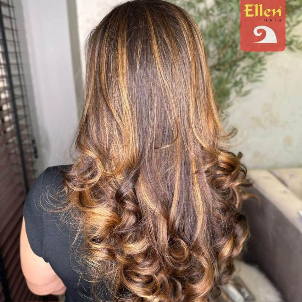 Mulher com cabelos castanhos longos e ondulados com mechas loiras. Ela veste uma camisa preta.