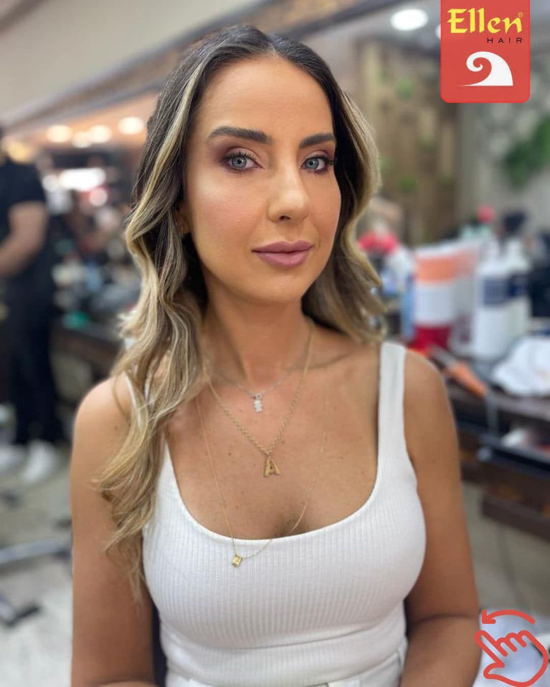 Mulher com cabelos loiros ondulados e maquiagem completa, usando uma regata branca em um salão de beleza.
