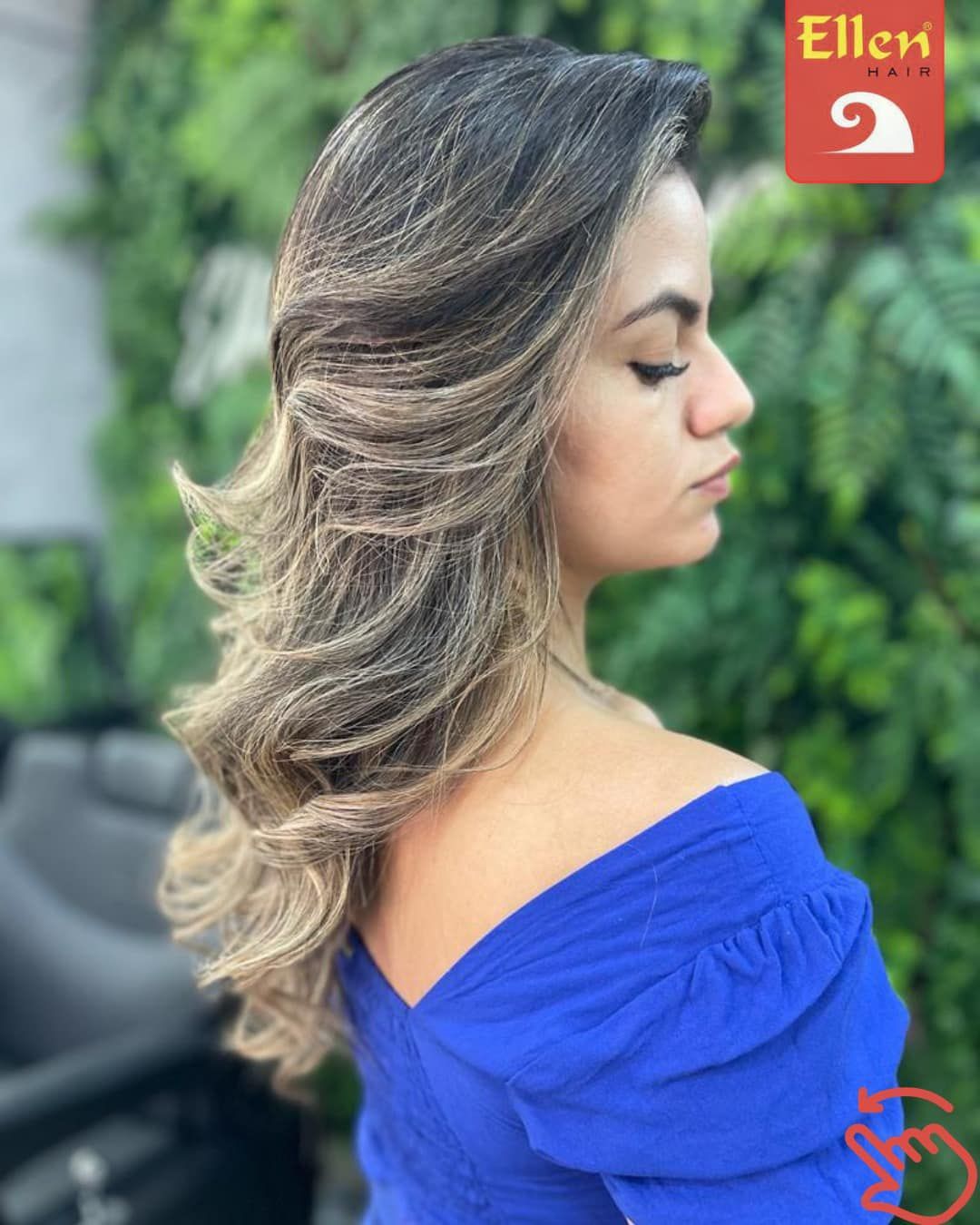 Mulher com cabelo ondulado em camadas e mechas, e blusa azul tomara que caia, posando em frente a uma parede verde.