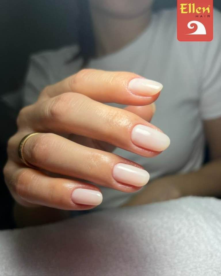 Uma mão com unhas ovais recém-feitas, de tom rosa-claro. Um anel de ouro no dedo anelar.