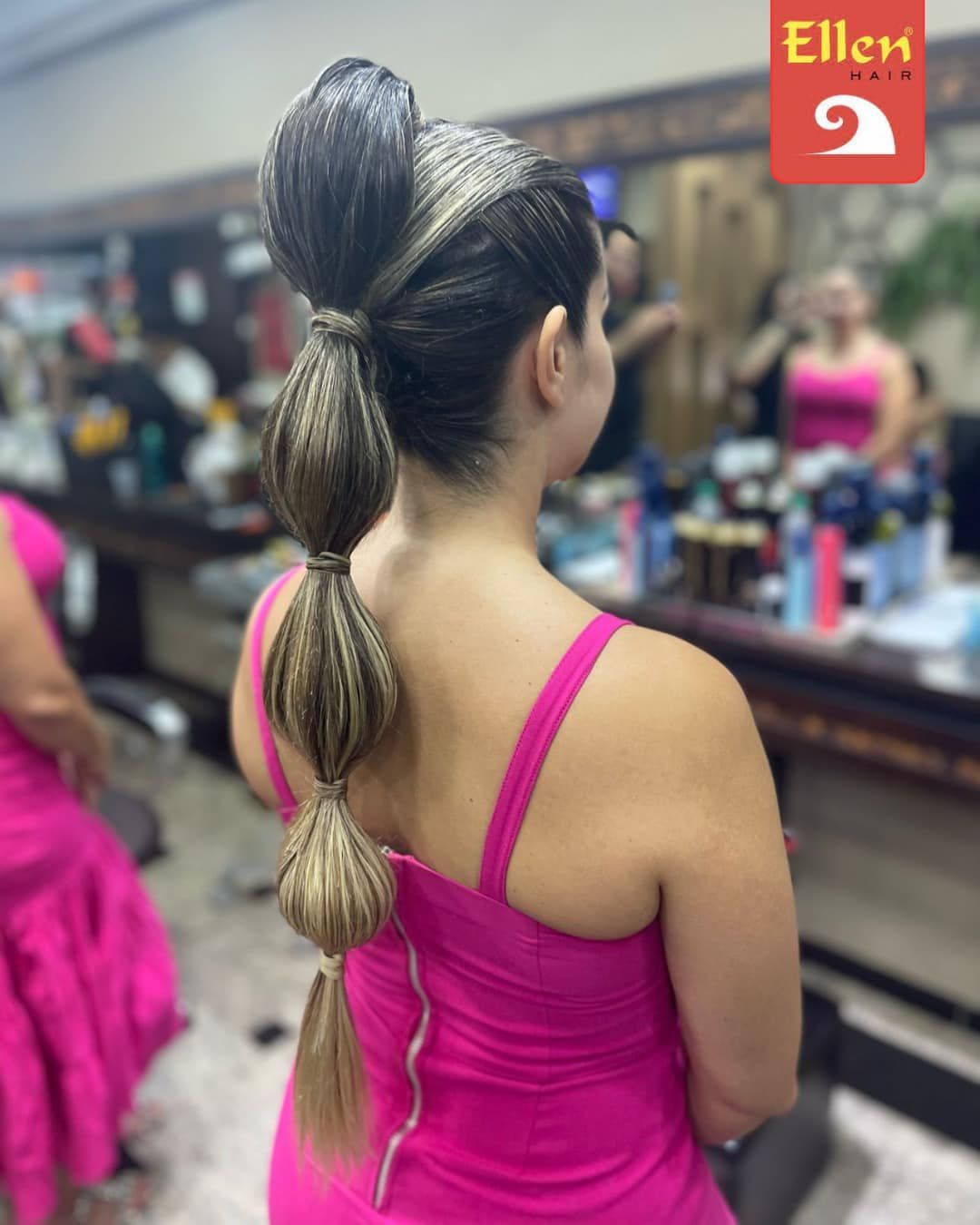 Mulher com rabo de cavalo trançado complexo em um salão, usando um vestido rosa. O cabelo está penteado com mechas bulbosas.