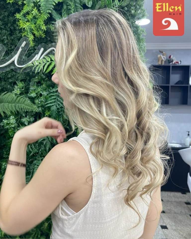 Mulher com cabelos loiros longos e ondulados no salão de beleza, posando com a mão perto do rosto. Parede verde ao fundo.