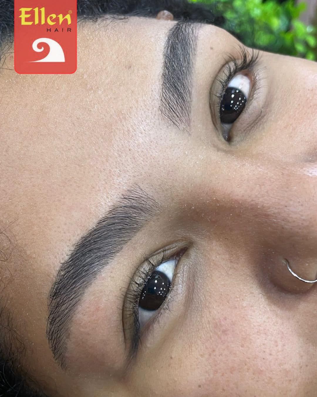 Close do rosto de uma pessoa com sobrancelhas escuras e preenchidas e um piercing no nariz. A pele é castanho-clara.