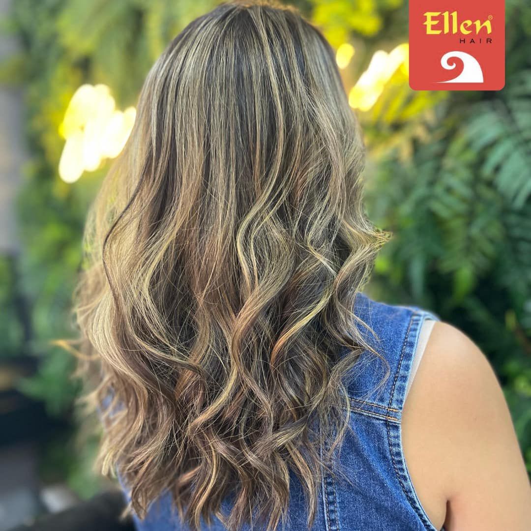 Mulher de cabelos castanhos ondulados e mechas loiras; veste um colete jeans. Ao fundo, parede verde.