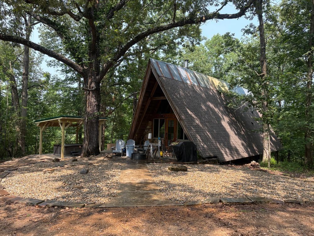 A-frame cabin in a wooded area; gravel yard, covered patio, tall trees, blue sky.