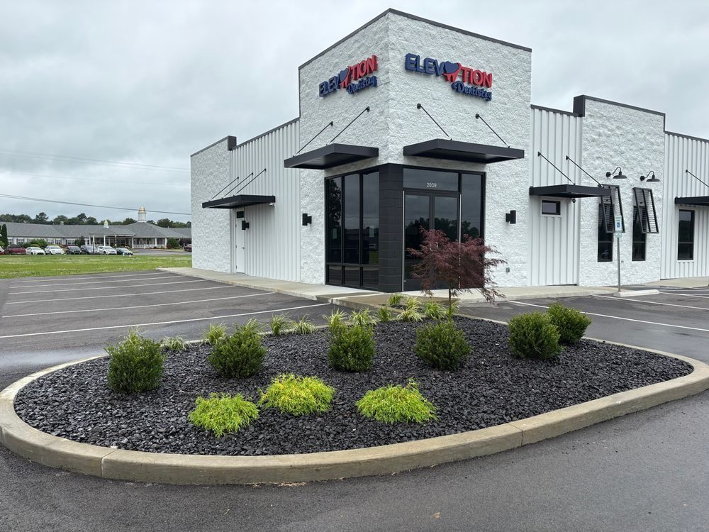 Modern white building with black trim, ELAVATION Fitness sign, landscaped bed with dark rocks and bushes.