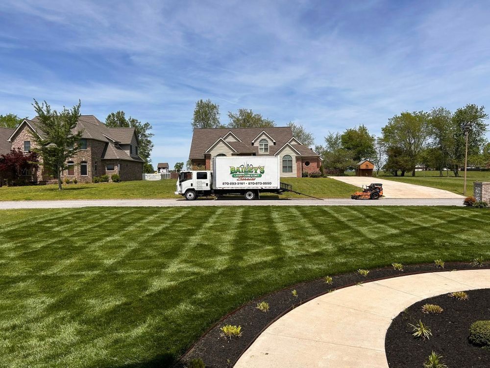 Lawn service truck and mower on a residential street. Green lawn with striped pattern, houses, and blue sky.