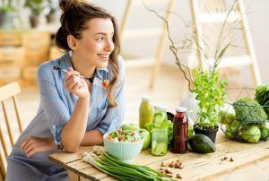 Woman smiles while eating salad at a wooden table with fresh produce and drinks.