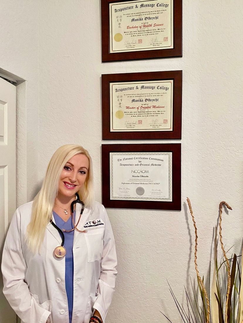 Woman in lab coat with stethoscope, smiling, stands near framed certificates on a wall.