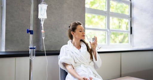 Woman in white robe receives IV drip, drinking water by a window.