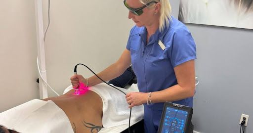 Person administering laser therapy on a patient's back in a clinic; bright pink light visible.