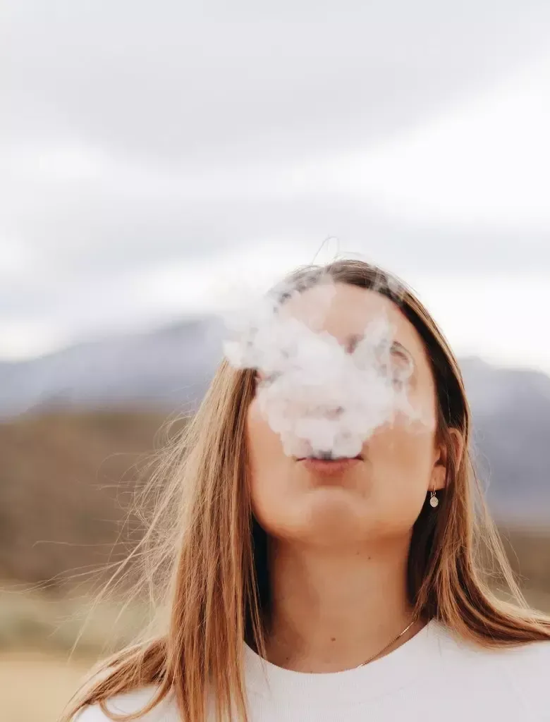 Woman exhales white vapor. Blonde hair, white shirt, outdoors, blurred mountain background.