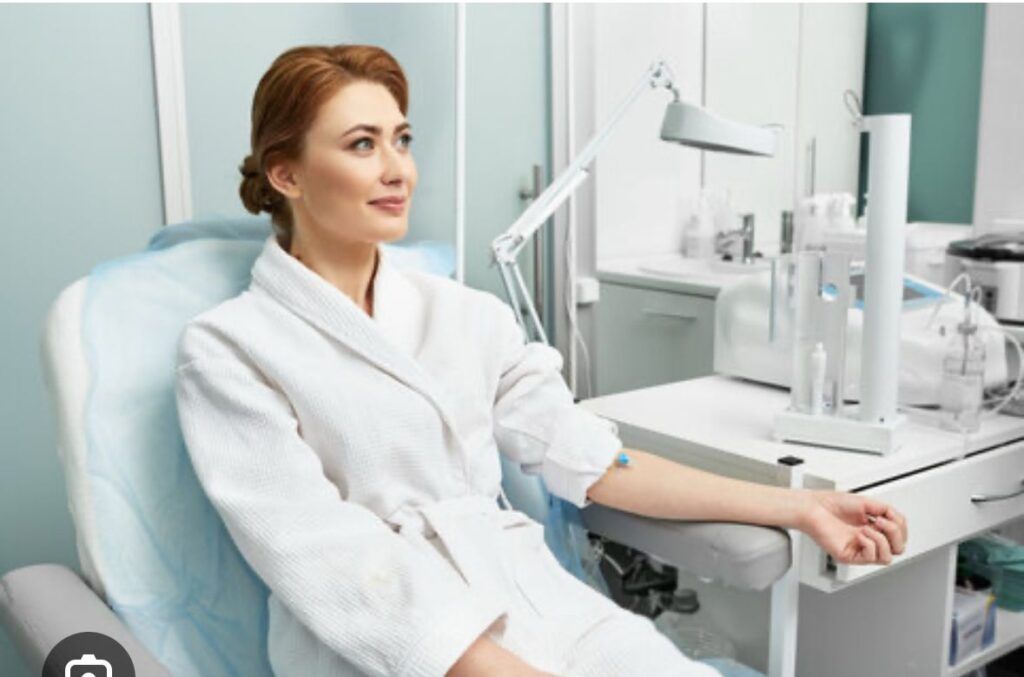 Woman in a white robe seated in a medical chair, arm extended with equipment nearby.