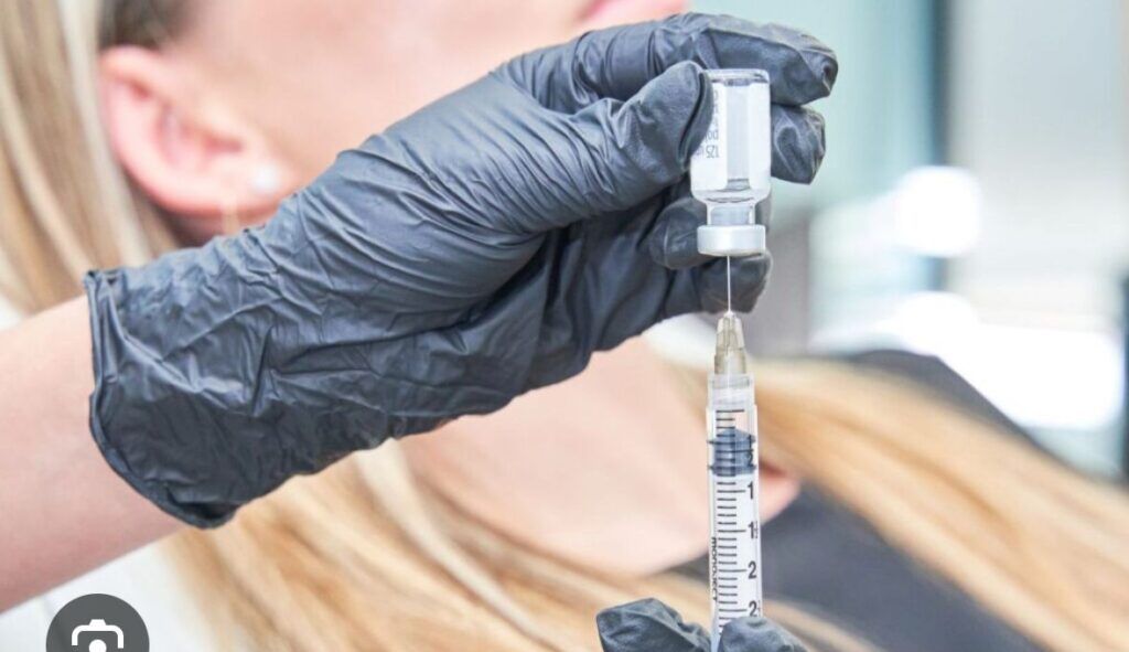 Gloved hand drawing liquid from a vial into a syringe. Blond hair and blurred background visible.