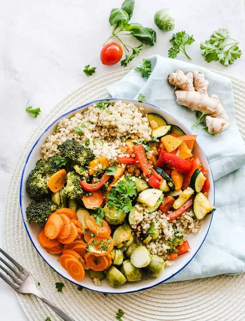Colorful vegetable and quinoa bowl with broccoli, carrots, peppers, Brussels sprouts, and herbs on a white plate.