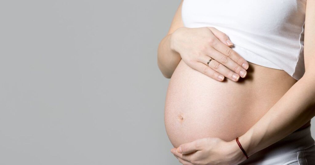 Pregnant person holding their belly, wearing a white top, gray background.