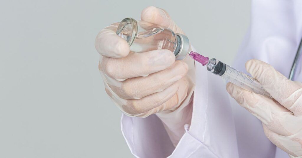 Gloved hands filling a syringe from a vaccine vial against a white backdrop.