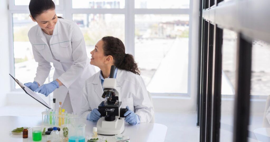 Two scientists in lab coats examine results near a microscope and colorful beakers in a bright laboratory.