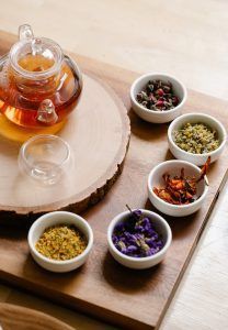 Glass teapot with tea and small bowls of dried herbs on a wooden board.