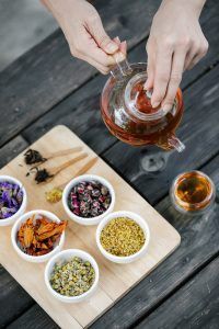 Person pouring tea from a glass teapot, wooden board with tea ingredients in small white bowls.