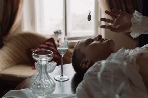 Person being hypnotized, lying down with a pendant swinging above them; a glass and decanter sit nearby.