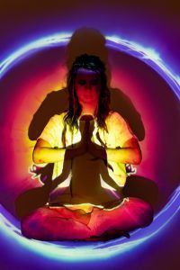 Person in yoga pose, hands together, lit with colorful aura.