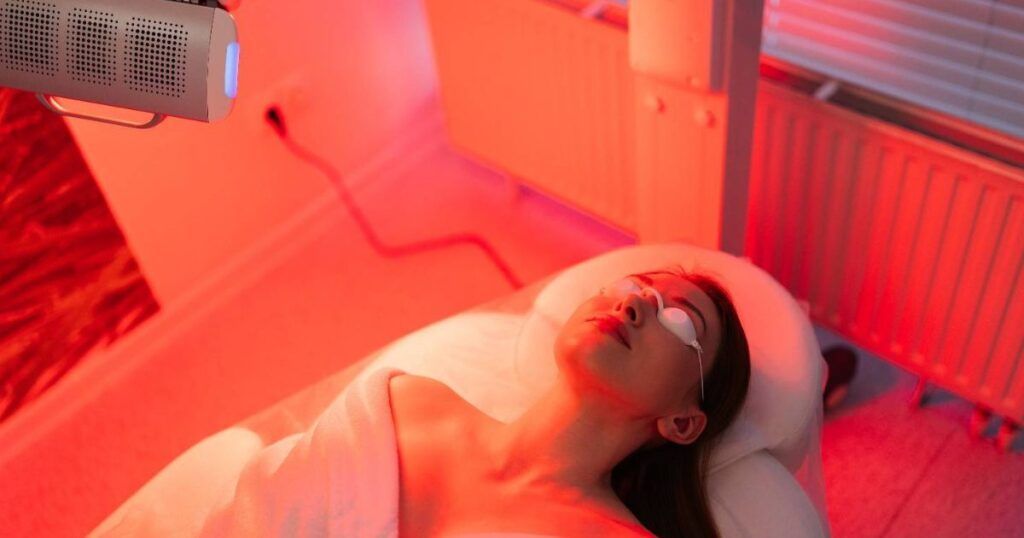 Woman with eye protection lies under red light therapy lamp in spa.