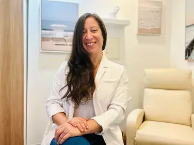 Woman in white coat smiles, sitting in an office with artwork on the wall.