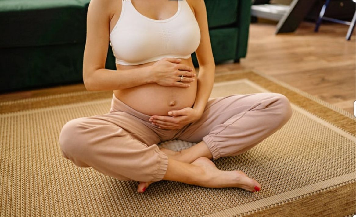 Pregnant person in a white top and tan pants sitting cross-legged on a rug, holding their belly.