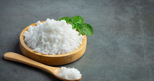 White salt in a wooden bowl with a wooden spoon on a gray surface, garnished with mint.