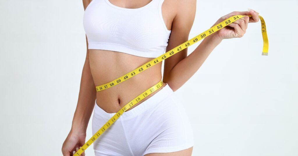 Woman in white activewear measures her waist with a yellow tape measure, white background.