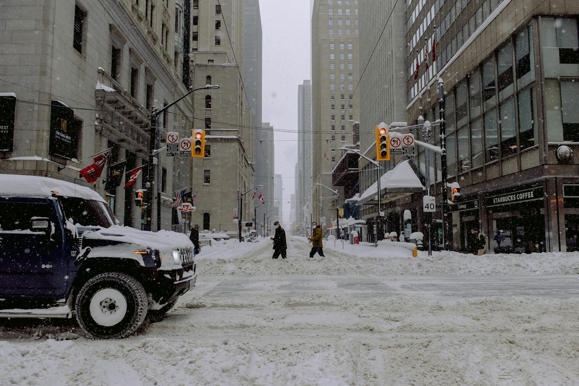 A snowy downtown street with a car driving in the foreground. Snow covers the road and sidewalks, wh