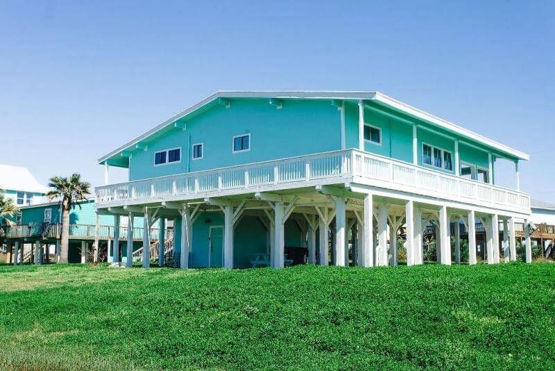 A large blue house with a white deck is sitting on top of a lush green field.