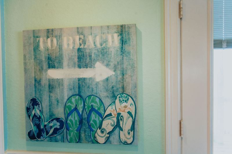 A painting of flip flops and an arrow pointing to the beach is hanging on a wall.